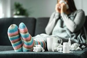 Sick woman with flu, cold, fever and cough sitting on couch at home. Ill person blowing nose and sneezing with tissue and handkerchief. Woolen socks and medicine. Infection in winter.